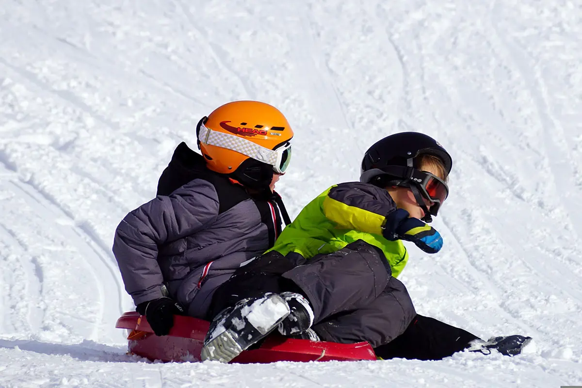 Twee kinderen sleeën op sneeuw met helmen en winteruitrusting op in Oostenrijk. Foto door Pixabay.