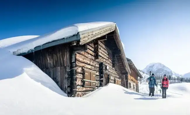 Zwei Menschen mit Skiausrüstung stehen vor einer verschneiten Hütte