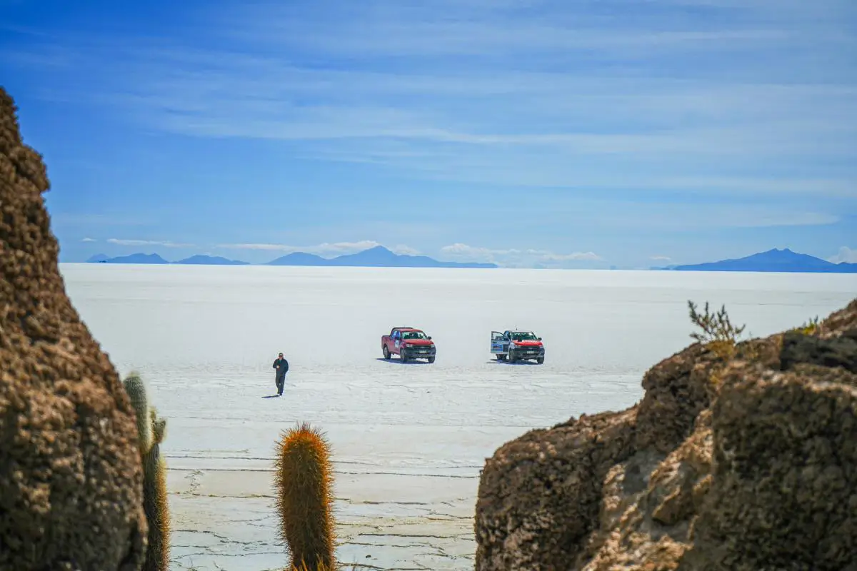 A person walks away from two cars parked on a salt flat in Bolivia towards a cactus filled island. Photo By Becca Marsh.