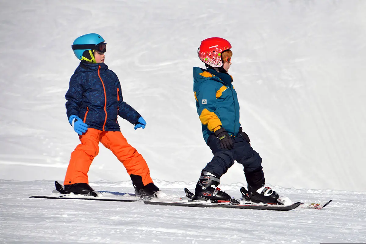 Zwei Kinder fahren mit Helmen und Winterkleidung auf einem verschneiten Hang in Österreich Ski. Foto von Piro, Pixabay.