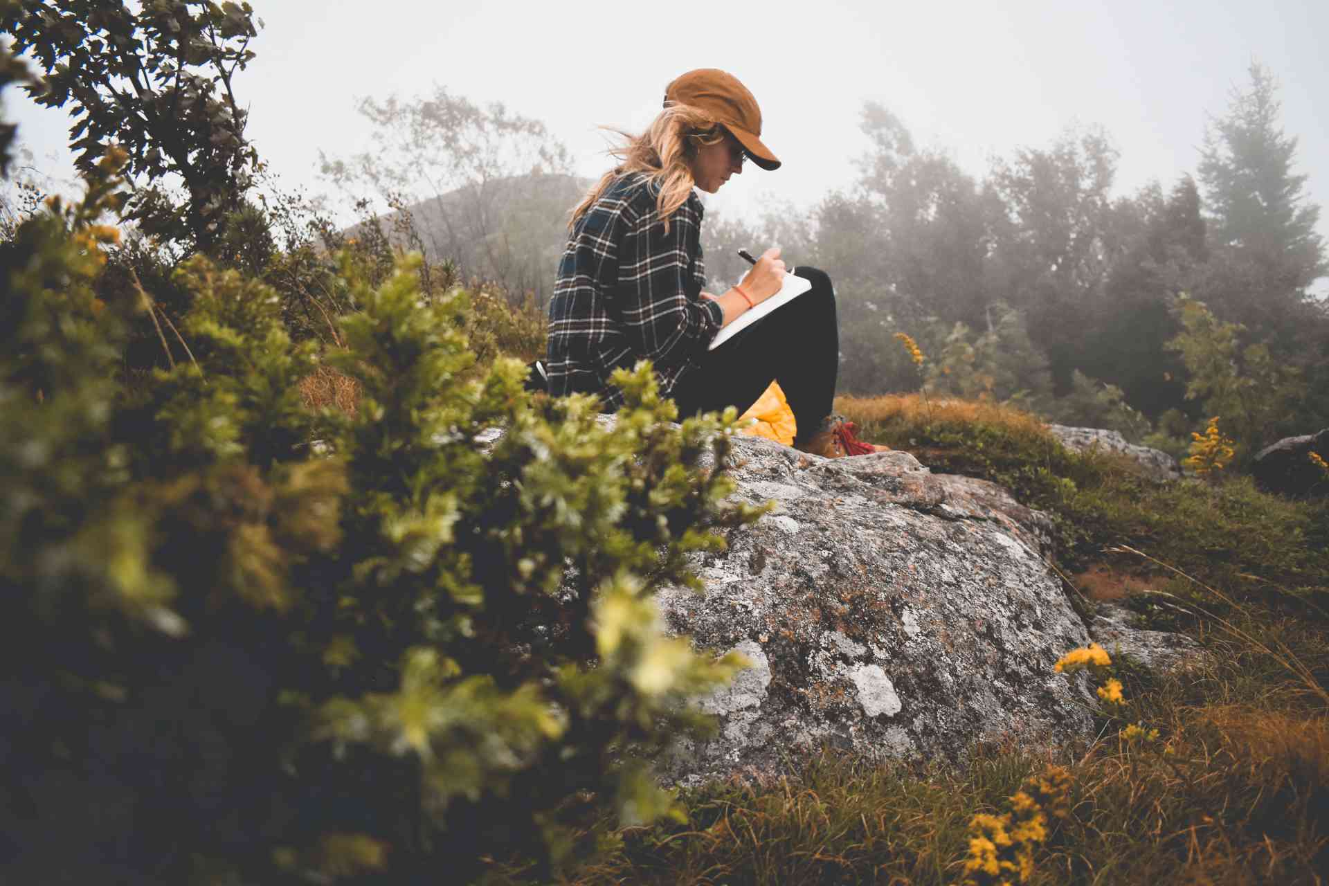 Hiker girl sitting on a rock in a forest is writing about adventure travel in her notebook