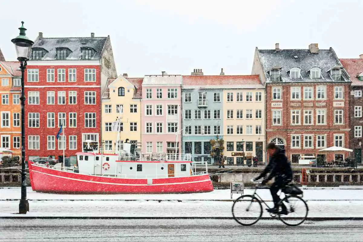 A person on a bike cycling past the colourful district of Nyhavn in Copenhagen on a snowy winters day. Photo by Max Adulyanukosol from Unsplash
